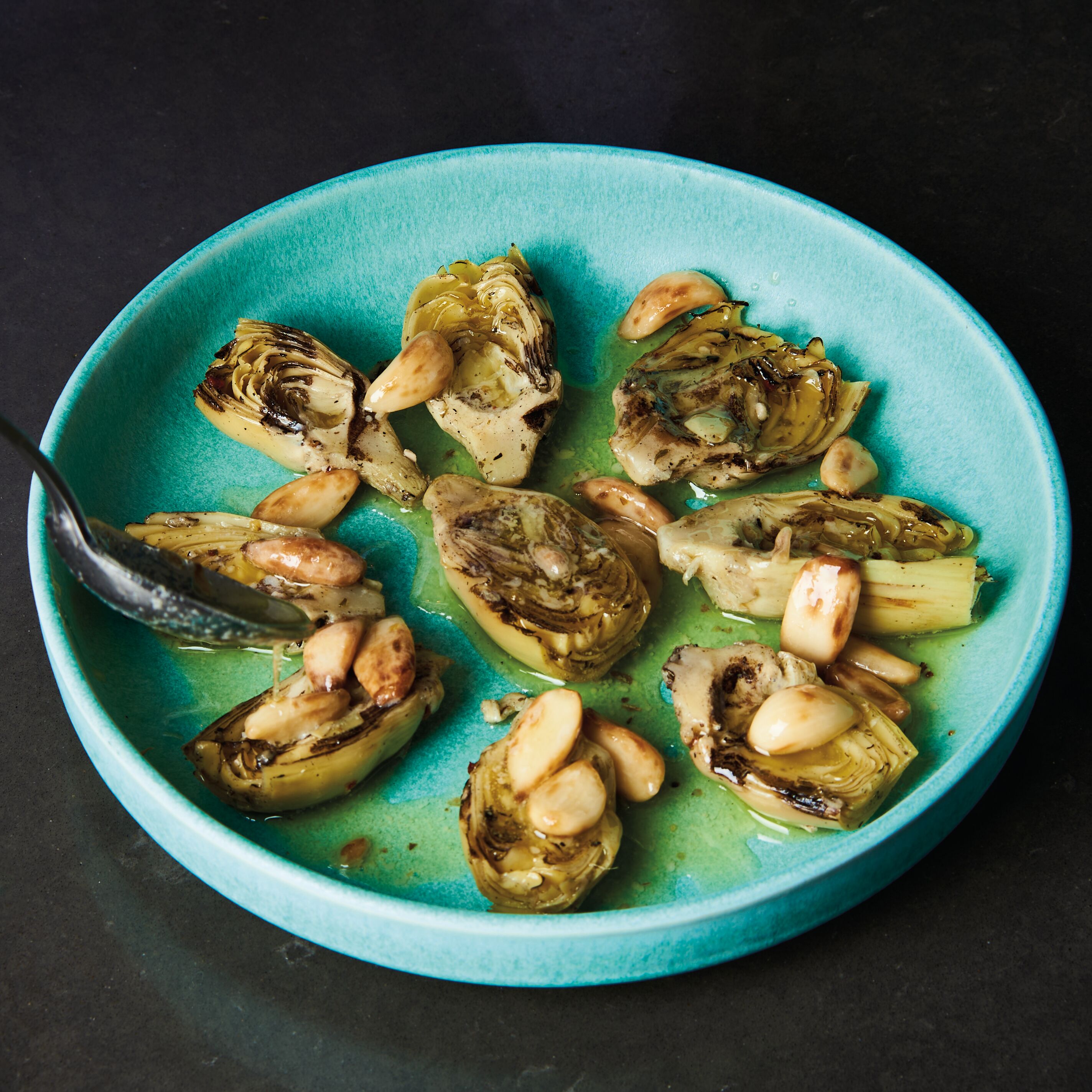 Grilled artichokes arranged on a blue plate with roasted garlic cloves and lemon garlic butter being drizzled with a spoon