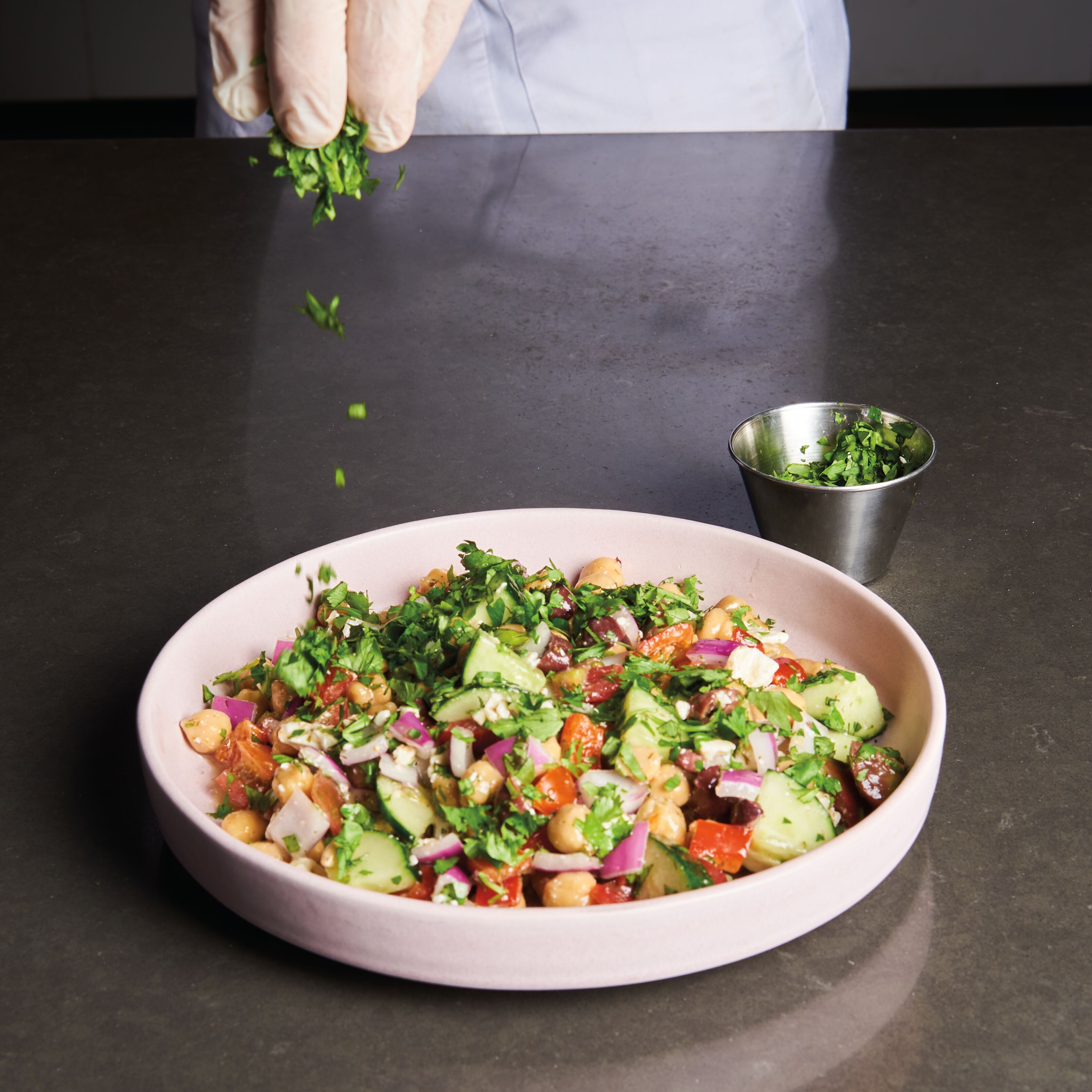 chef's hand sprinkling parsley over a salad of chickpeas, cucumbers, red onion, tomato, and kalamata olives in a pink bowl