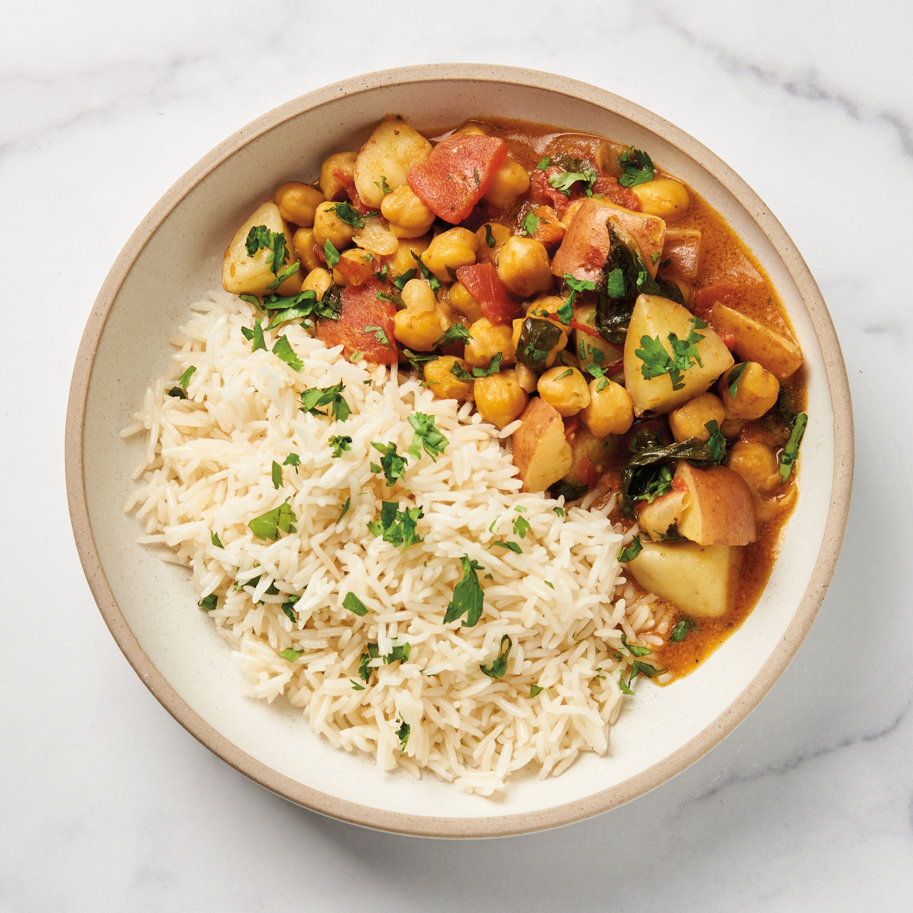 Curry made from chickpeas, potatoes, and tomatoes in a white bowl with basmati rice and cilantro garnish