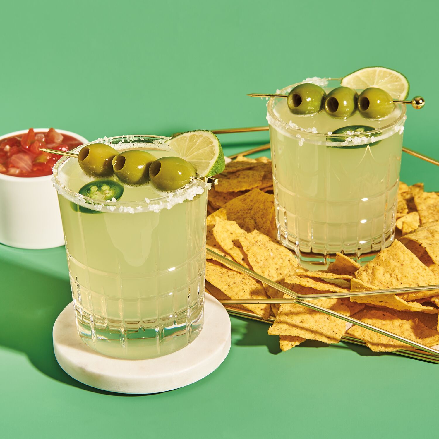 two margaritas topped in glasses rimmed with salt, topped with jalapeno, lime, and olive garnish with chips and salsa in background